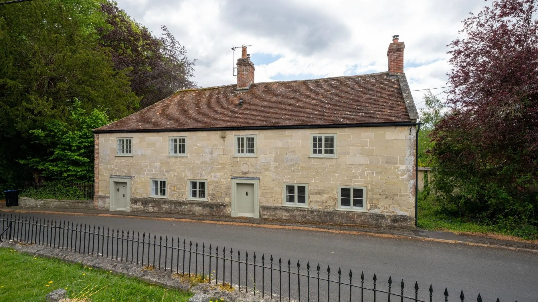 Double-fronted pale stone house with sage-green window frames and a clay tile roof beside a village lane