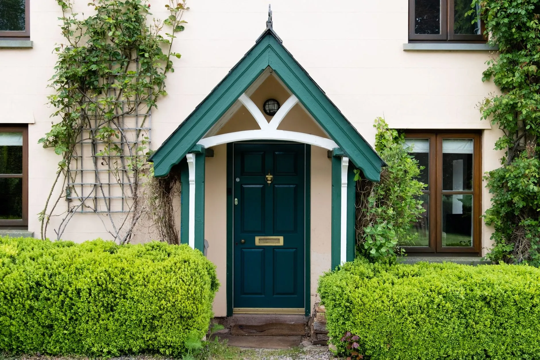Cream house front with a green gabled wooden porch over a dark green door, between box hedges